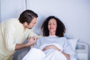 Patient assis sur un lit d’hôpital, une main posée sur le bas-ventre pour évoquer une douleur liée à une sonde JJ.